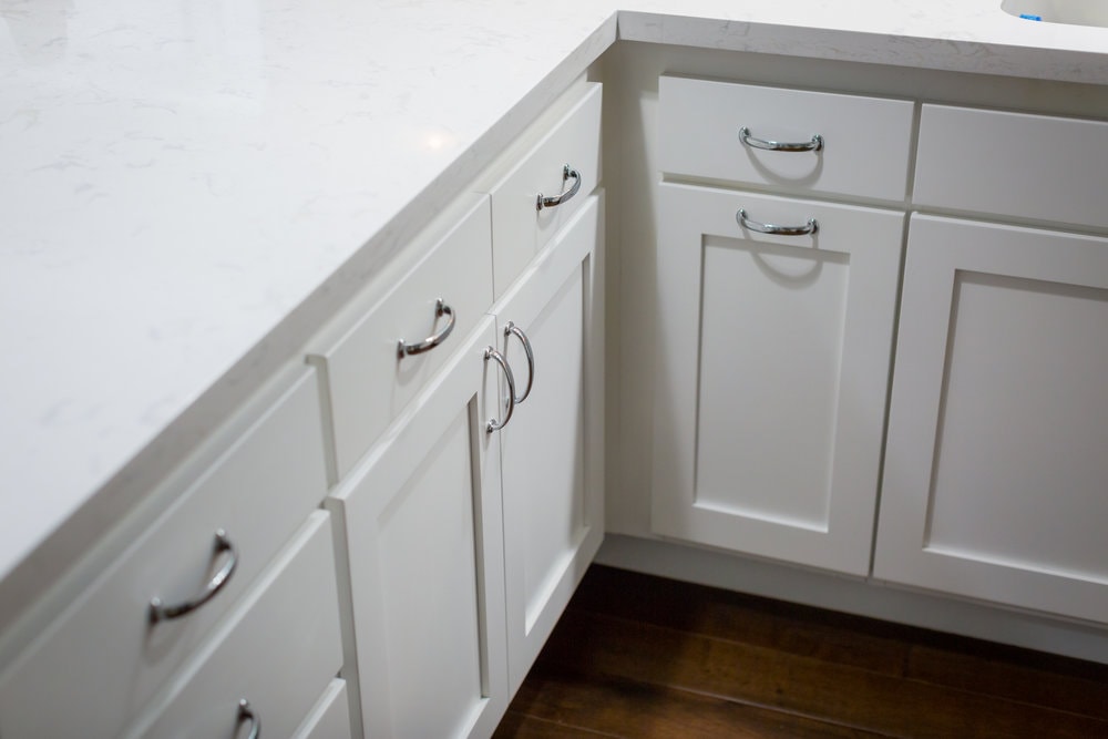 White kitchen cabinets with silver handles and a white marble countertop on a dark wood floor.