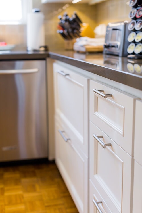 Close-up of modern white kitchen cabinets with stainless steel handles and a dishwasher on the left.
