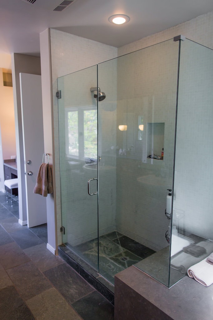 Modern glass-enclosed shower with tiled walls and floor, next to a towel rack and open bathroom door.