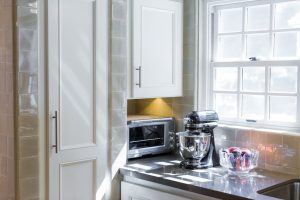Modern kitchen corner with a stand mixer, microwave, bowl of fruit, white cabinets, and sunlight through a window.