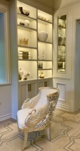 Elegant white and gold chair with winged arms sits before a modern lit shelf, perfect for a cabinet refresh.
