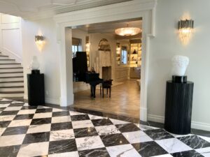 Elegant foyer with black and white checkered marble floor, sculpted busts on black pedestals, wall sconces, and an open archway leading to a room with a grand piano and bookshelves. Curved staircase on the left.